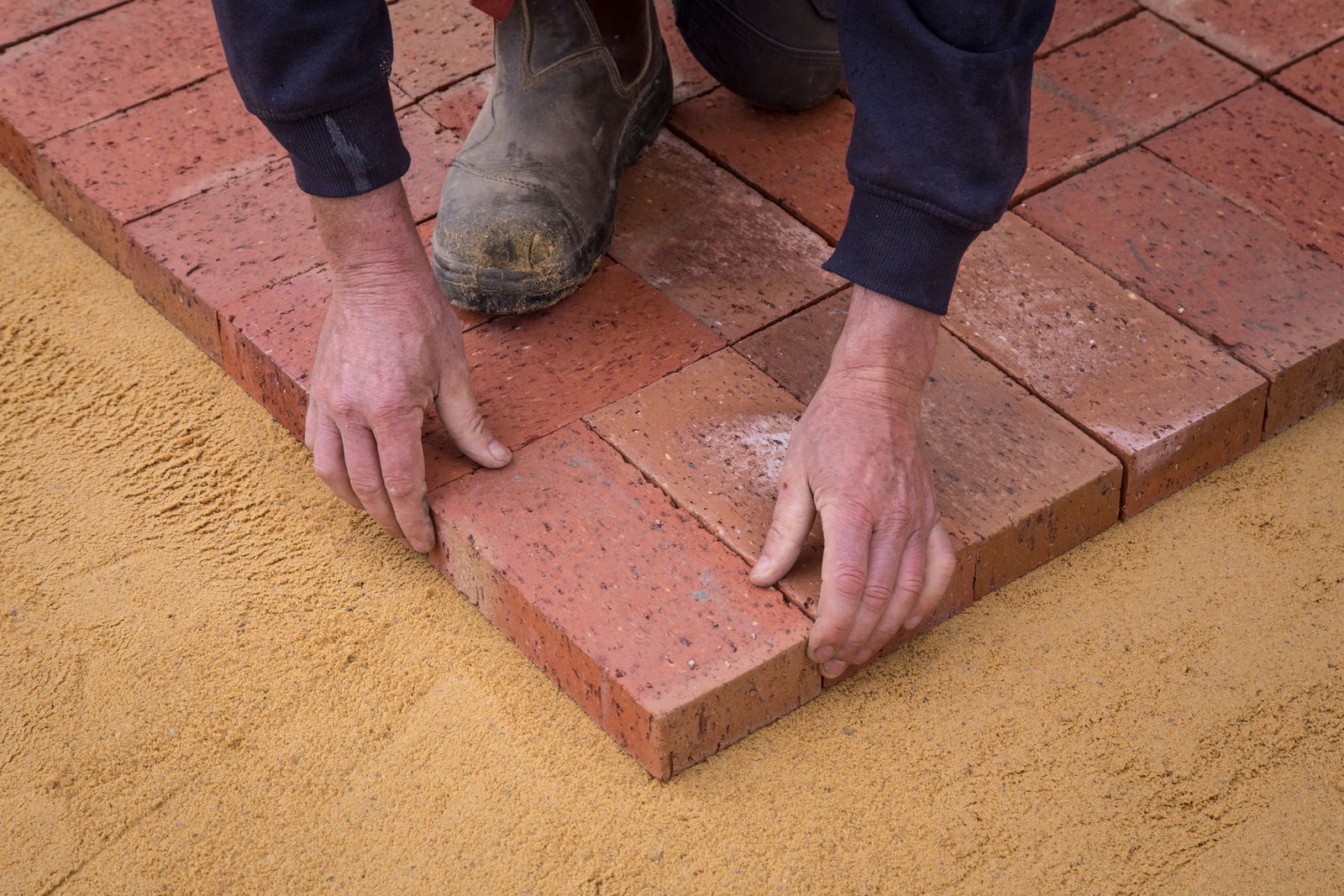 Hands setting a clay paver into a leveled sand bed during a walkway installation
